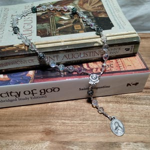 May include: A white and green rosary with clear glass beads and silver accents. The rosary is resting on top of two books, one titled "The Confessions of Saint Augustine" and the other titled "The City of God (Abridged Study Edition)".