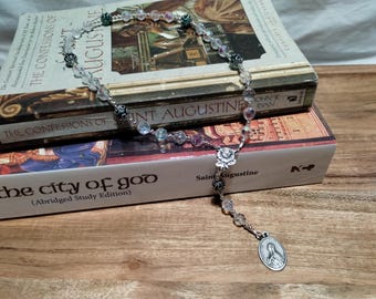 Op de afbeelding: Een wit en groene rozenkrans met heldere glazen kralen en zilveren accenten. De rozenkrans ligt op twee boeken, een met de titel "De Belijdenissen van Sint Augustinus" en de andere met de titel "De stad van God (Korte editie)".