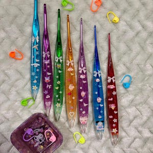 Pode incluir: Uma cole&ccedil;&atilde;o de agulhas de croch&ecirc; coloridas com glitter e desenhos de personagens. As agulhas s&atilde;o em tons de azul, roxo, verde, laranja e vermelho. Tamb&eacute;m est&atilde;o inclu&iacute;dos v&aacute;rios marcadores de ponto e um pequeno prato quadrado.