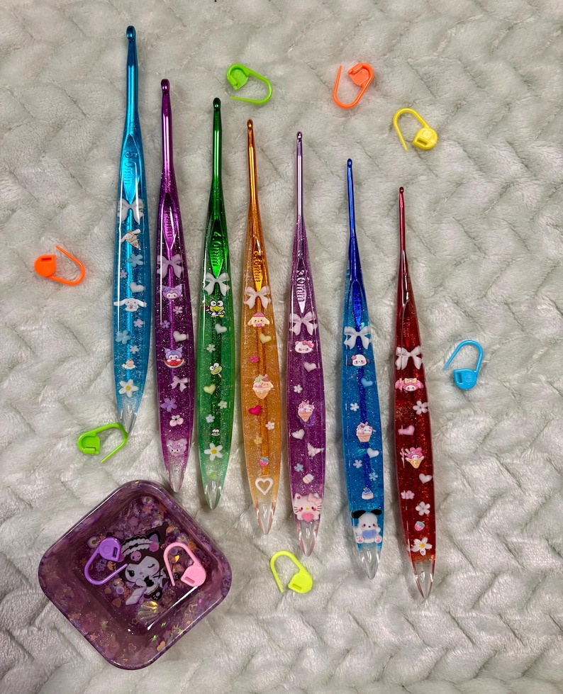 Pode incluir: Uma cole&ccedil;&atilde;o de agulhas de croch&ecirc; coloridas com glitter e desenhos de personagens. As agulhas s&atilde;o em tons de azul, roxo, verde, laranja e vermelho. Tamb&eacute;m est&atilde;o inclu&iacute;dos v&aacute;rios marcadores de ponto e um pequeno prato quadrado.
