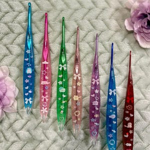 May include: A collection of seven colorful crochet hooks. Each hook features a clear, glitter-filled handle with decorative elements like bows, stars, and clouds. The hooks come in various colors, including blue, pink, green, and purple, and are arranged on a textured surface.