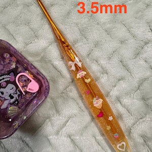 Pode incluir: Um gancho de croch&ecirc; dourado com um cabo transparente preenchido com elementos decorativos, incluindo um cupcake, cora&ccedil;&otilde;es e la&ccedil;os. O gancho tem 3,5 mm.