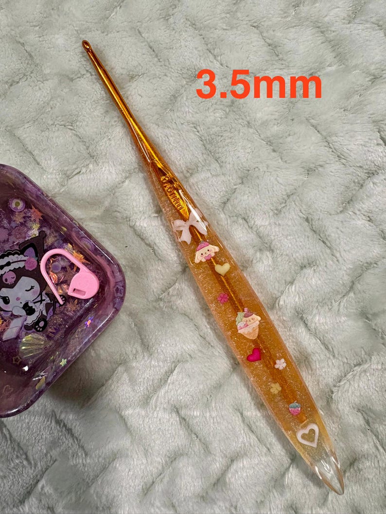 Pode incluir: Um gancho de croch&ecirc; dourado com um cabo transparente preenchido com elementos decorativos, incluindo um cupcake, cora&ccedil;&otilde;es e la&ccedil;os. O gancho tem 3,5 mm.
