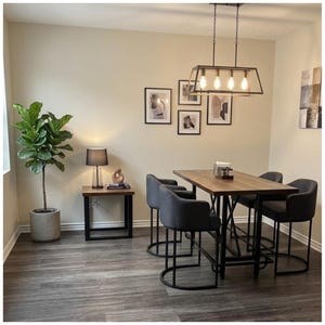 May include: A dining room set featuring a rectangular wooden table with a black metal frame and four matching dark gray upholstered chairs. A black metal pendant light hangs above the table, and a potted fiddle leaf fig plant stands nearby.