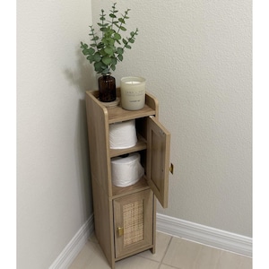 May include: A light wood-tone bathroom storage cabinet with a small shelf, two open compartments for toilet paper, and a lower cabinet with a woven panel. A candle and a vase with greenery sit on top. The cabinet is placed in a corner.