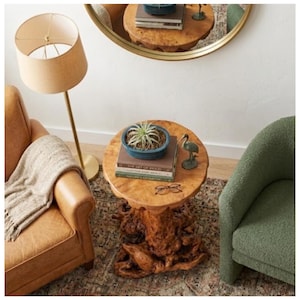 May include: A round wooden side table with a unique root base. The table holds a blue ceramic pot with a plant, books, and a small bird figurine. A tan lamp and leather chair are in the background.