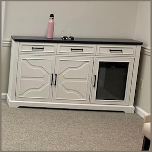 May include: A white cabinet with a black countertop. The cabinet has three drawers and three doors, with a glass-fronted section. A pink water bottle and sunglasses rest on top.