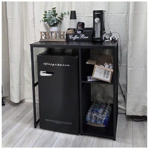 May include: A black coffee bar with a mini-fridge, shelving, and a countertop. The countertop holds a coffee maker, cups, and a sign that says "Coffee Bar". A small plant and the word "FAITH" are also on the countertop. The mini-fridge has the Frigidaire logo.