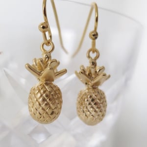 Könnte beinhalten: Goldene Ananas-Ohrringe mit strukturierter Oberfläche. Die Ohrringe befinden sich auf einem weißen Hintergrund.