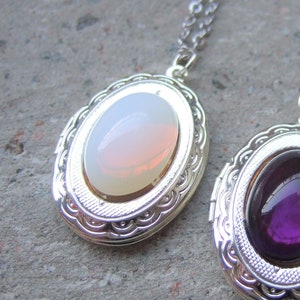 May include: Two silver locket necklaces with oval-shaped glass inserts. One locket has a white and pink iridescent stone, the other has a purple stone.