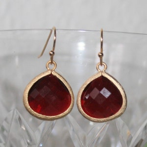 May include: A pair of teardrop-shaped earrings with faceted, deep red gems set in gold-colored metal. The earrings hang from delicate gold-colored hooks, showcasing a classic and elegant design.