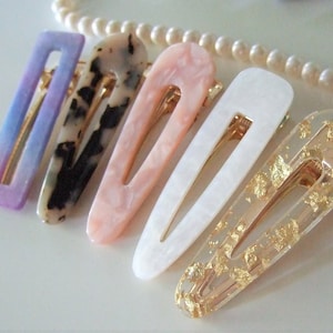 Può includere: Sei diverse mollette per capelli con vari motivi e colori. Le mollette sono in plastica e hanno una molla in metallo dorato. I motivi includono un ombré viola, un motivo marmoreo bianco e nero, un motivo marmoreo rosa, un motivo marmoreo bianco e una molletta trasparente con scaglie dorate.