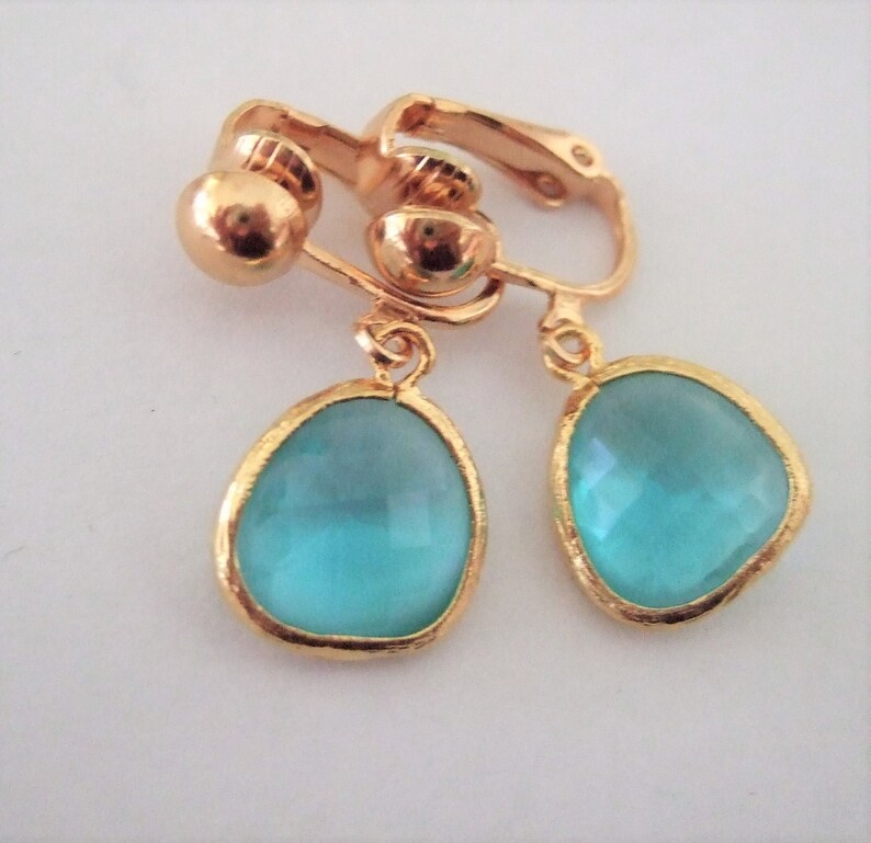 Puede incluir: Un par de pendientes de clip de tono dorado con piedras de vidrio azul turquesa en forma de l&aacute;grima.