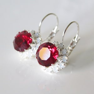May include: A pair of leverback earrings with large, faceted, ruby-red gemstones encircled by smaller, white, opalescent stones. The earrings have a silver-coloured metal setting, displayed on a white surface.