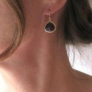 May include: A close-up of a gold-colored earring with a teardrop-shaped, dark purple stone. The earring is hanging from a woman's ear. The woman has light skin and blonde hair.