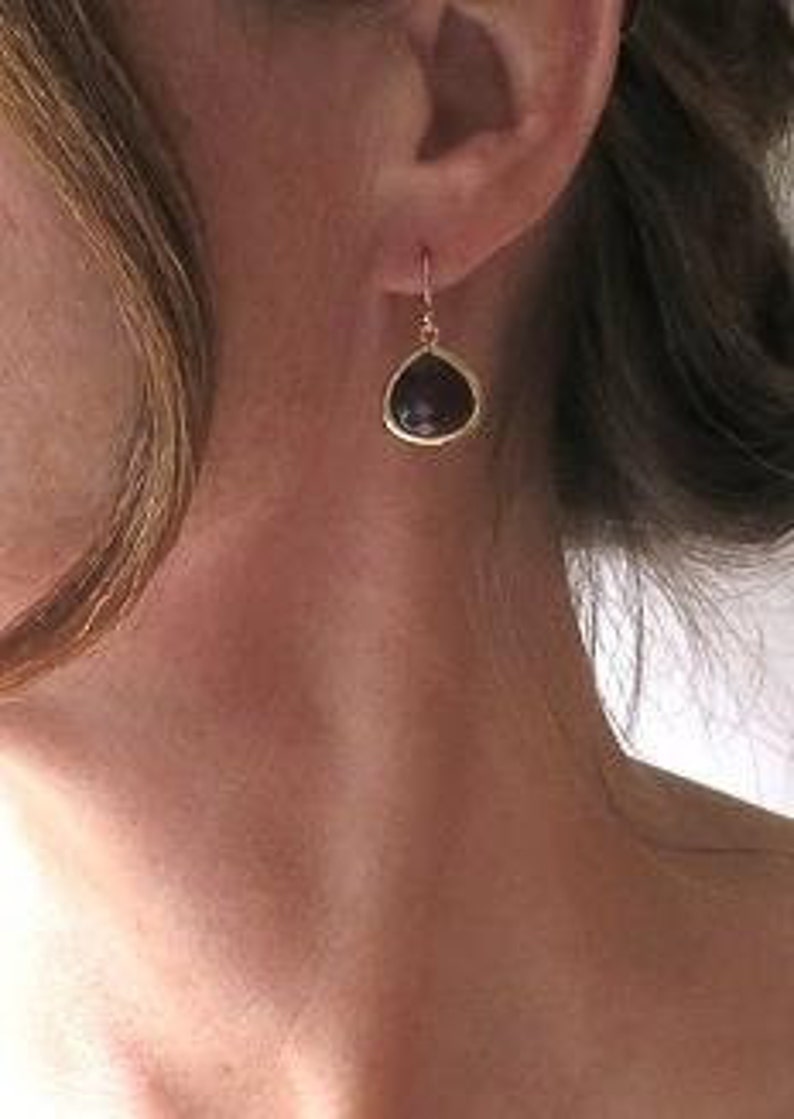 May include: A close-up of a gold-colored earring with a teardrop-shaped, dark purple stone. The earring is hanging from a woman's ear. The woman has light skin and blonde hair.