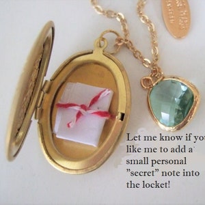 May include: Gold locket necklace with a small white note inside and a green gemstone pendant. The locket is open and the note is visible. The text on the image reads: "Let me know if you like me to add a small personal "secret" note into the locket!"