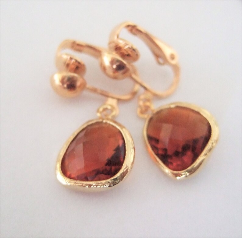 Puede incluir: Un par de pendientes de clip de tono dorado con piedras de vidrio de color &aacute;mbar en forma de l&aacute;grima.