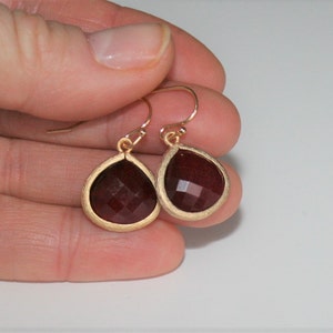 May include: A pair of gold-colored drop earrings with teardrop-shaped, faceted, dark red gemstones. The gemstones are set in a gold-colored bezel. The earrings are held in a person's hand against a white background.