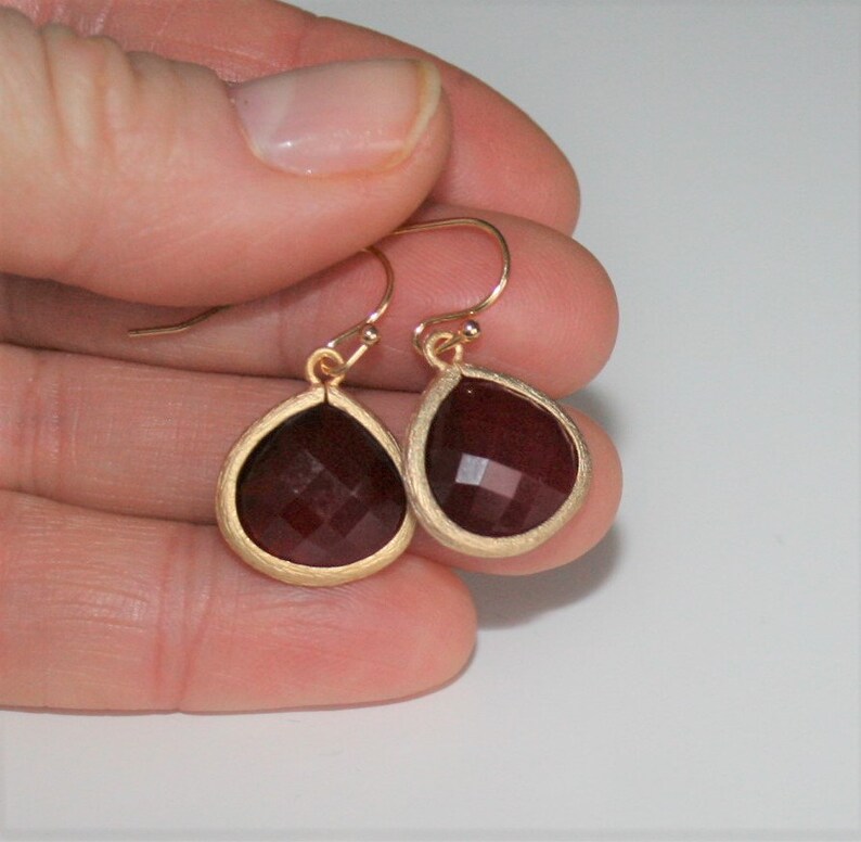May include: A pair of gold-colored drop earrings with teardrop-shaped, faceted, dark red gemstones. The gemstones are set in a gold-colored bezel. The earrings are held in a person's hand against a white background.