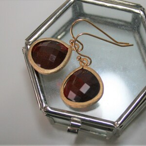 May include: A pair of gold-toned earrings with teardrop-shaped, faceted, dark red gemstones. The earrings are set in a gold-colored bezel and have a simple hook design. The earrings are displayed inside a clear, octagonal glass box.