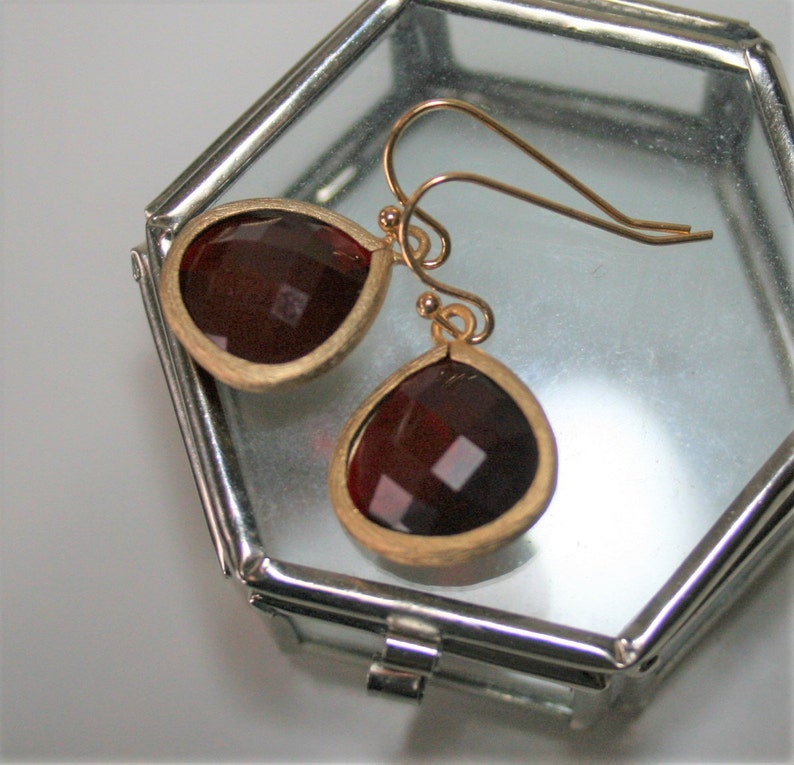 May include: A pair of gold-toned earrings with teardrop-shaped, faceted, dark red gemstones. The earrings are set in a gold-colored bezel and have a simple hook design. The earrings are displayed inside a clear, octagonal glass box.