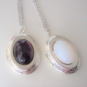 May include: Two silver locket necklaces with oval-shaped glass inserts. One locket has a dark purple glass insert and the other has a white glass insert.