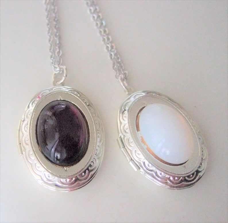 May include: Two silver locket necklaces with oval-shaped glass inserts. One locket has a dark purple glass insert and the other has a white glass insert.