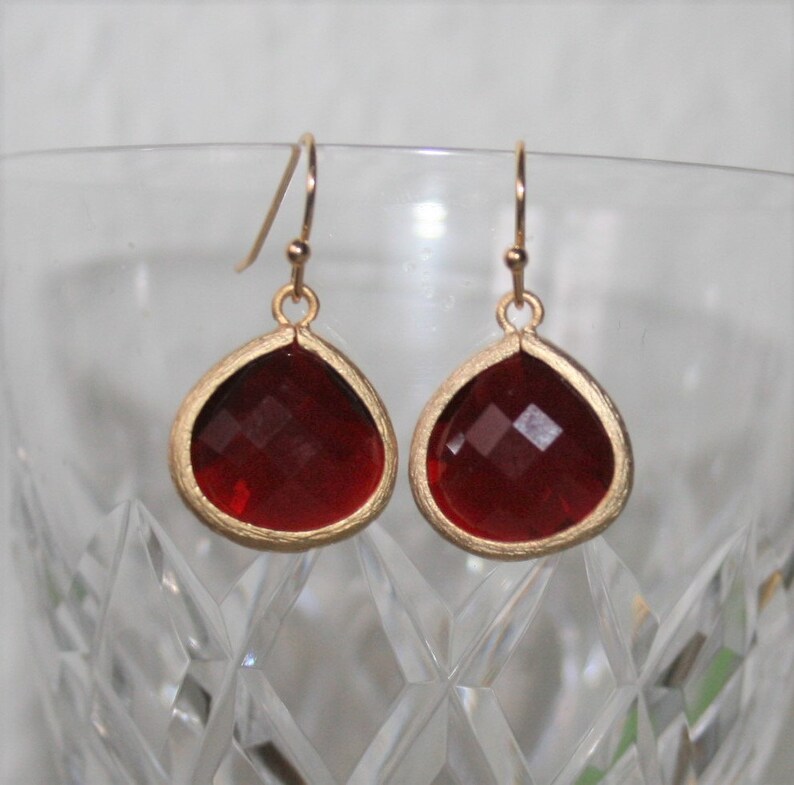 May include: A pair of teardrop-shaped earrings with faceted, deep red gems set in gold-colored metal. The earrings hang from delicate gold-colored hooks, showcasing a classic and elegant design.