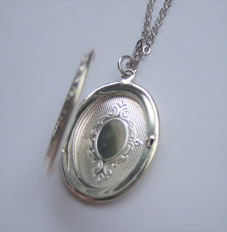 May include: Silver oval locket necklace with a detailed engraved design on the front. The locket is open, revealing a blank interior.