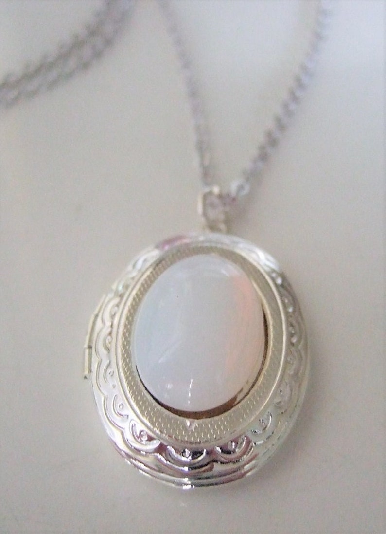 May include: A silver locket necklace with a white oval-shaped stone in the center. The locket has a detailed, ornate design around the edge.