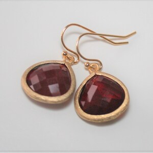 May include: A pair of gold-colored earrings with teardrop-shaped, faceted, dark red gemstones. The gemstones are set in a textured gold-colored metal. The earrings have a simple hook design.