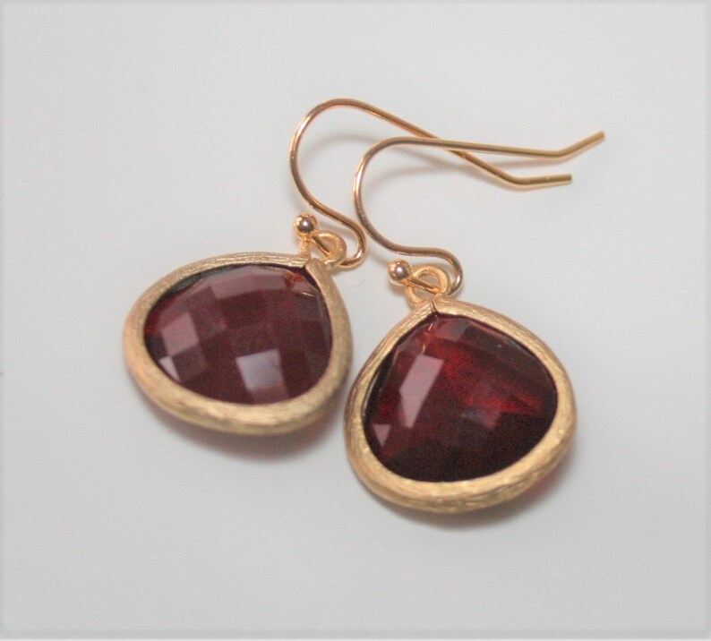 May include: A pair of gold-colored earrings with teardrop-shaped, faceted, dark red gemstones. The gemstones are set in a textured gold-colored metal. The earrings have a simple hook design.