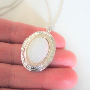 May include: A silver oval locket necklace with a white stone in the center. The locket has a detailed design around the edge.