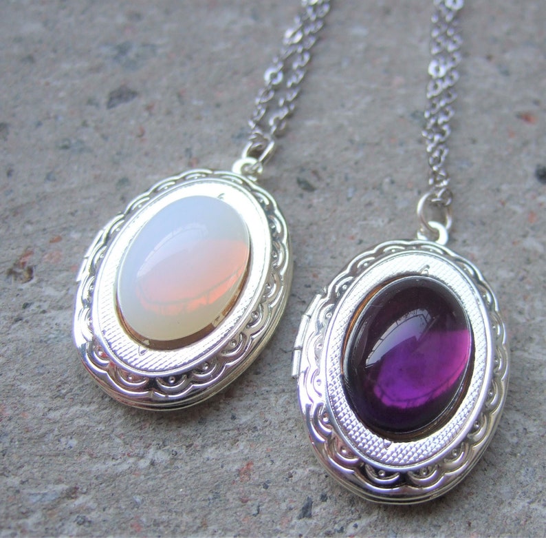 May include: Two silver locket necklaces with oval-shaped glass inserts. One locket has a white and pink iridescent stone, the other has a purple stone.