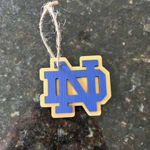 May include: Wooden ornament featuring the interlocking letters "ND" in blue, set against a gold-colored border. A natural twine loop is attached for hanging. The ornament is likely a decorative item or souvenir.