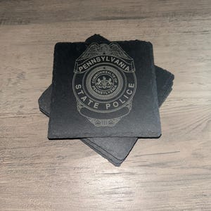 May include: Set of four black slate coasters with a laser-etched design of the Pennsylvania State Police badge. The badge features the words "Pennsylvania State Police" and the Commonwealth of Pennsylvania seal.