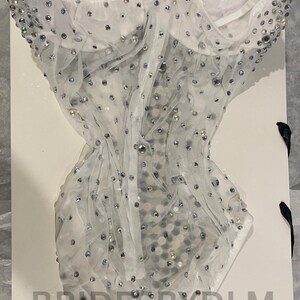 May include: A white, strapless bodysuit with a sheer overlay, embellished with silver sequins and pearls. The garment is displayed on a white surface, with the text "BRIDESBYDLM" visible.