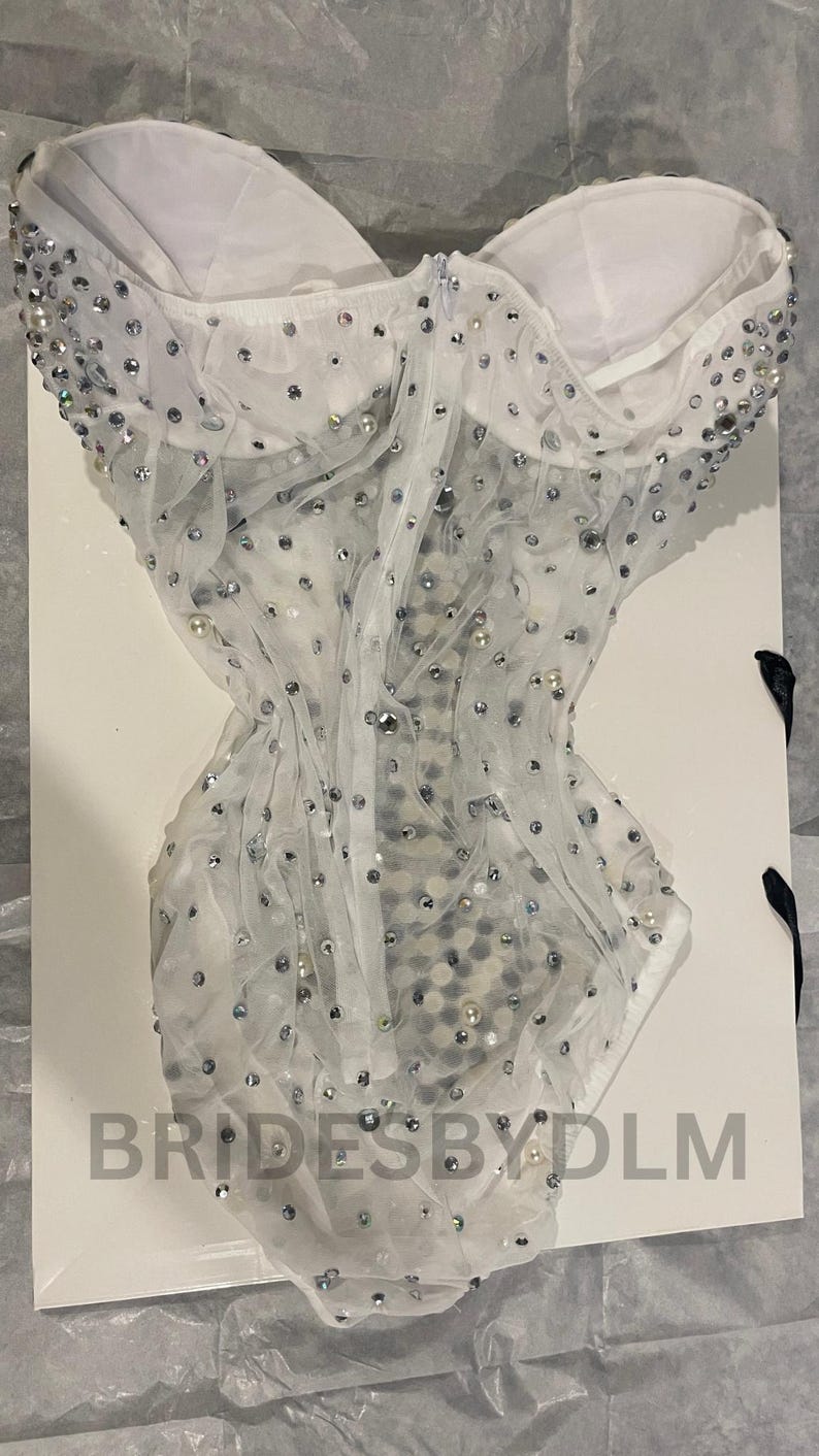May include: A white, strapless bodysuit with a sheer overlay, embellished with silver sequins and pearls. The garment is displayed on a white surface, with the text "BRIDESBYDLM" visible.
