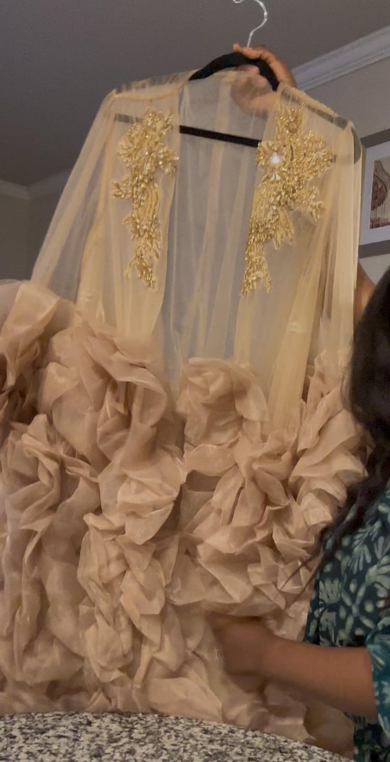 May include: A beige, sheer cape dress with gold embellishments. The cape features intricate gold embroidery on the shoulders. The lower portion of the dress has a layered, ruffled design in a similar beige hue, creating a voluminous effect. The dress is held up by a black hanger.