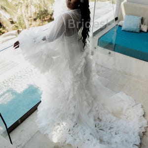 May include: A white bridal robe with a long train. The robe features layers of ruffled fabric, creating a voluminous effect. The sheer sleeves and bodice are visible. The bride is wearing a headpiece with a sparkling design. The background includes a pool and tropical foliage.