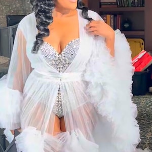 May include: A white, sheer, floor-length robe with ruffled trim and a matching belt. The robe is worn over a silver, jewel-encrusted bodysuit. The model has dark hair and is wearing a jeweled hair accessory.