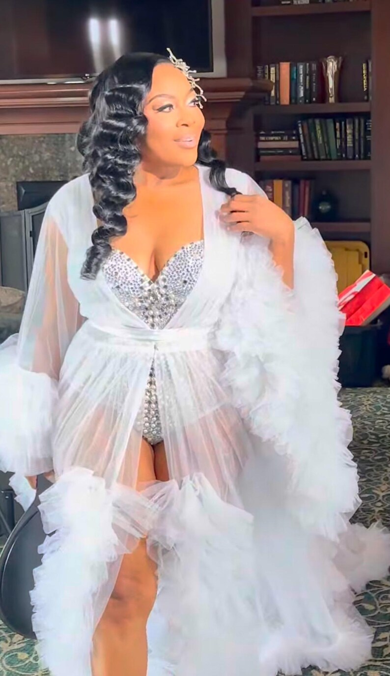 May include: A white, sheer, floor-length robe with ruffled trim and a matching belt. The robe is worn over a silver, jewel-encrusted bodysuit. The model has dark hair and is wearing a jeweled hair accessory.