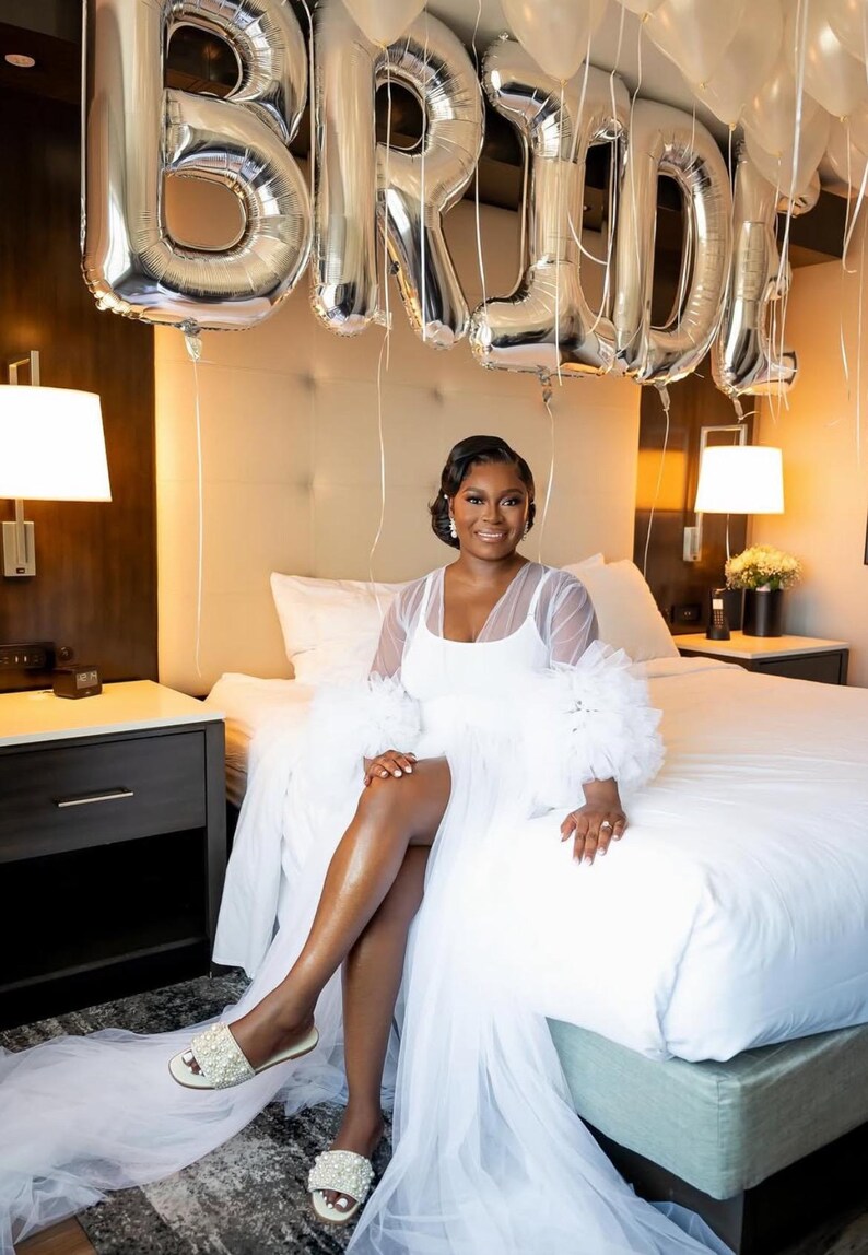 May include: A bride wearing a white tulle robe with a white satin slip dress underneath. The bride is sitting on a white bed with silver balloons that spell out 'BRIDE' hanging above the bed.