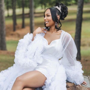 May include: A white tulle robe with a ruffled trim and long sleeves. The robe has a sweetheart neckline and a satin bodice. The robe is worn over a white satin mini dress.