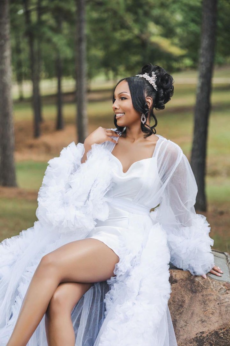 May include: A white tulle robe with a ruffled trim and long sleeves. The robe has a sweetheart neckline and a satin bodice. The robe is worn over a white satin mini dress.