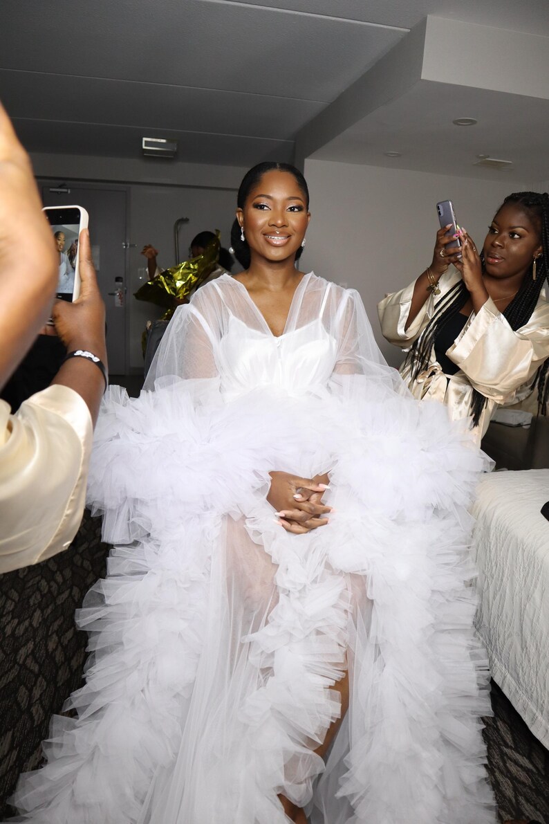 May include: A white tulle dress with a ruffled skirt and long sleeves. The dress is sheer and has a fitted bodice. The person is wearing a white dress with a ruffled skirt and long sleeves.