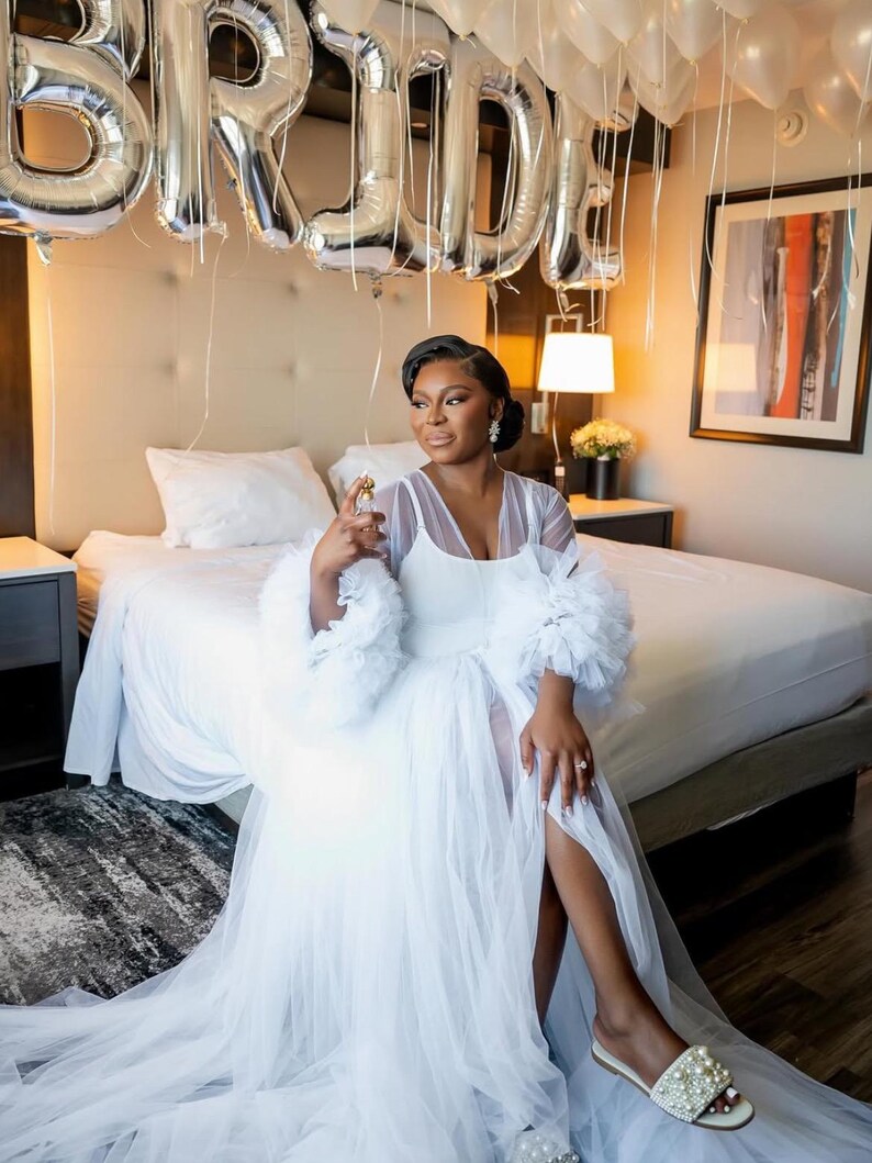 May include: A white tulle bridal robe with feather trim on the sleeves. The robe has a long train and a slit in the front. The bride is wearing white pearl slippers.