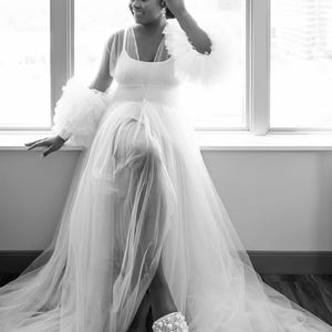 May include: A woman is wearing a white tulle gown with a sheer top and ruffled sleeves. She is wearing white pearl embellished sandals.
