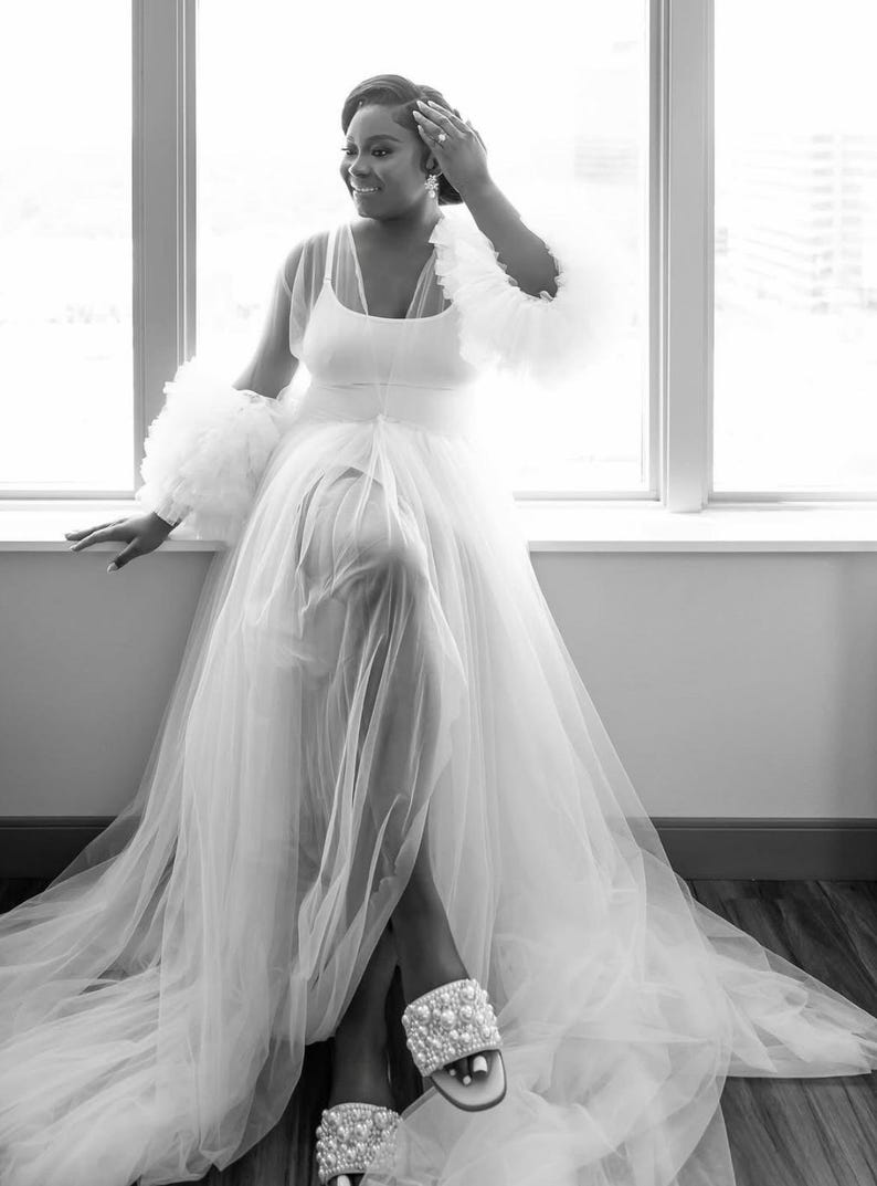 May include: A woman is wearing a white tulle gown with a sheer top and ruffled sleeves. She is wearing white pearl embellished sandals.